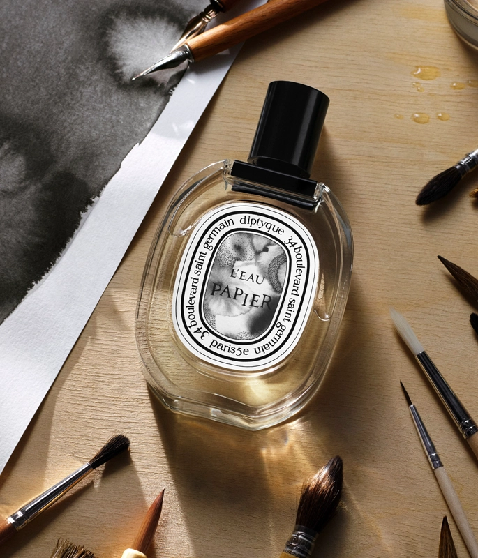 Diptyque perfume bottle flat lay surrounded by paint brushes and paintings 
